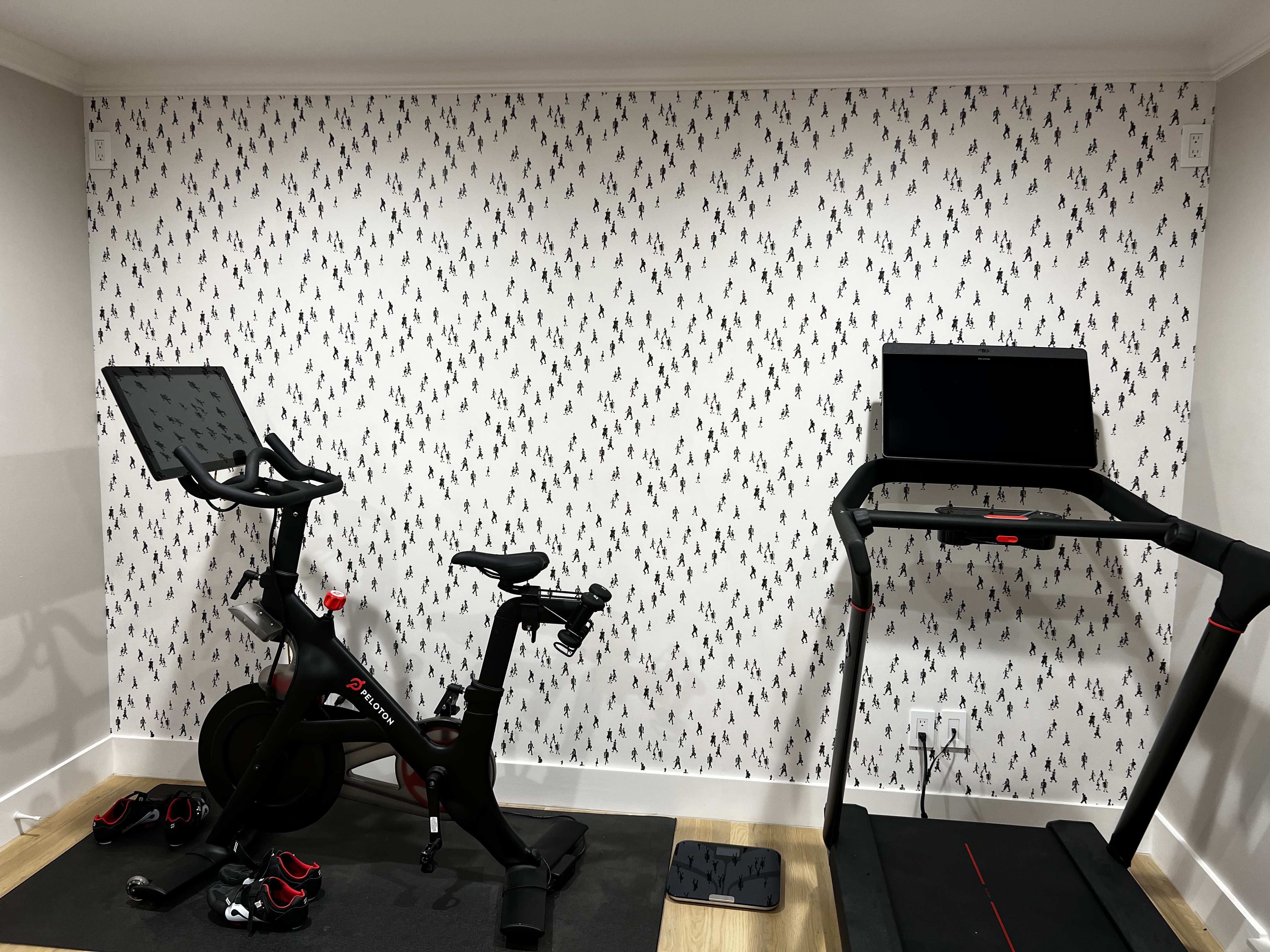 Home gym with playful walking-figures wallpaper behind a Peloton bike and treadmill