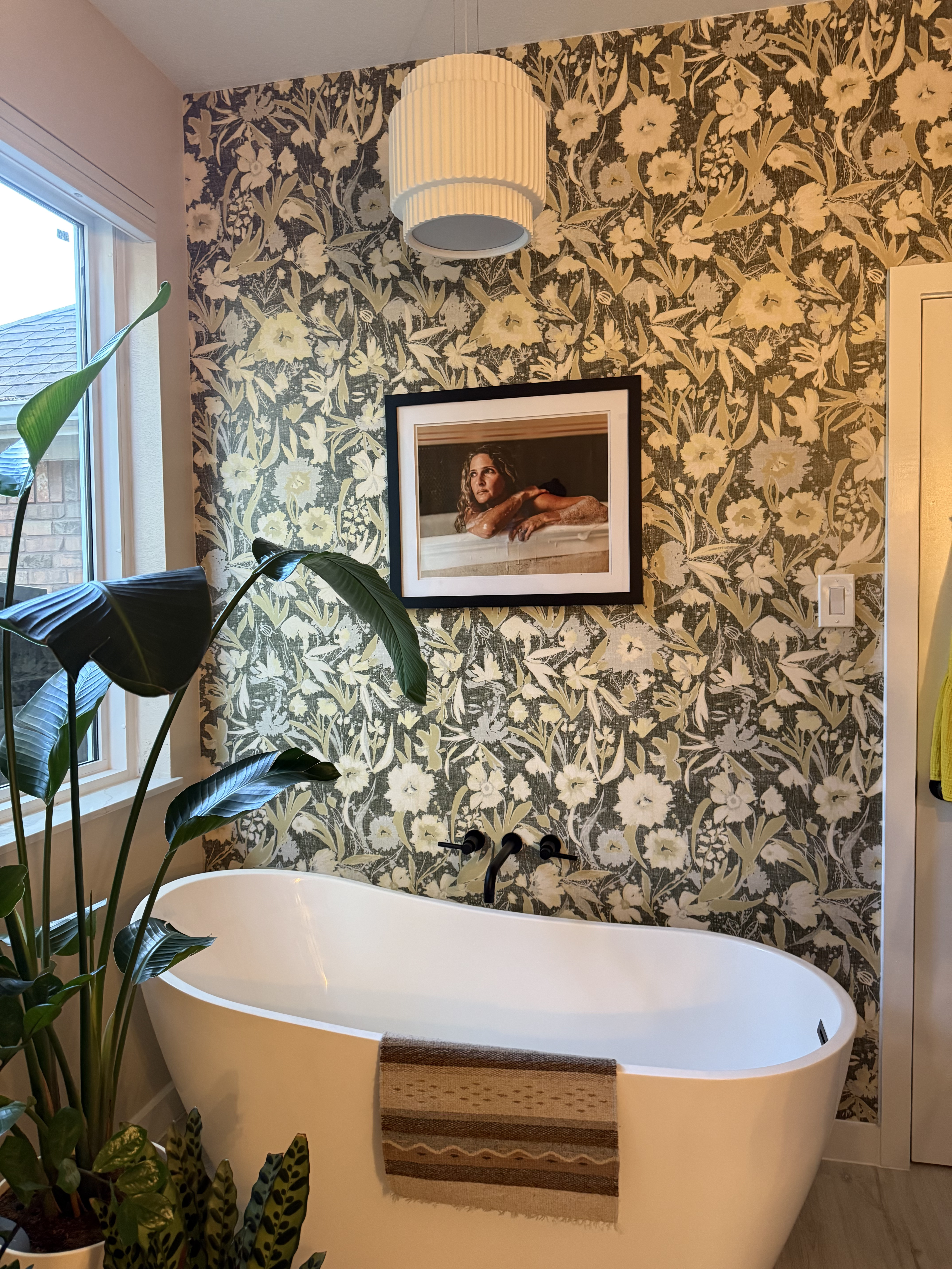 Freestanding tub against botanical wallpaper with pendant light and bird-of-paradise plant