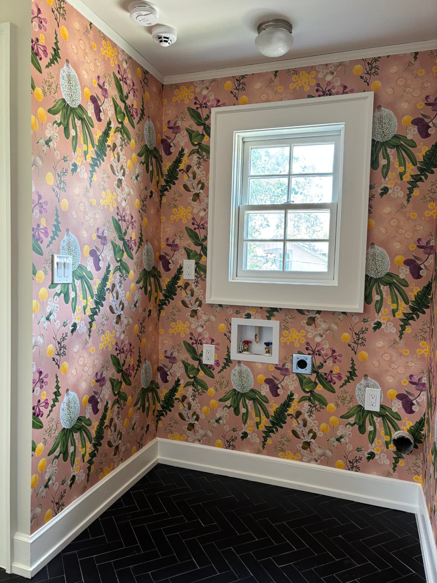 Laundry room in blush botanical wallpaper with white trim and black herringbone tile floor