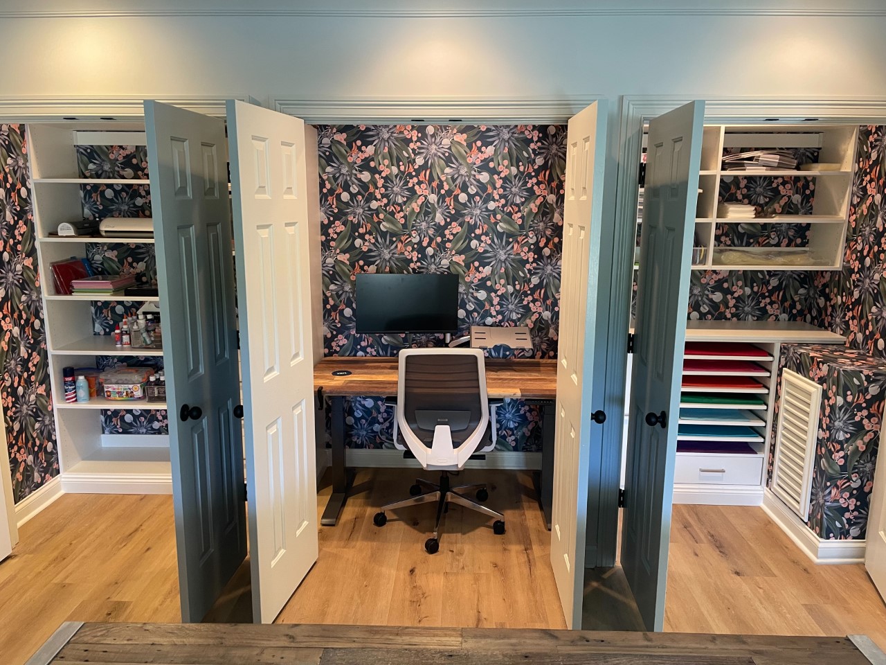 Craft closet lined in dark floral wallpaper with built-in desk and colorful paper storage