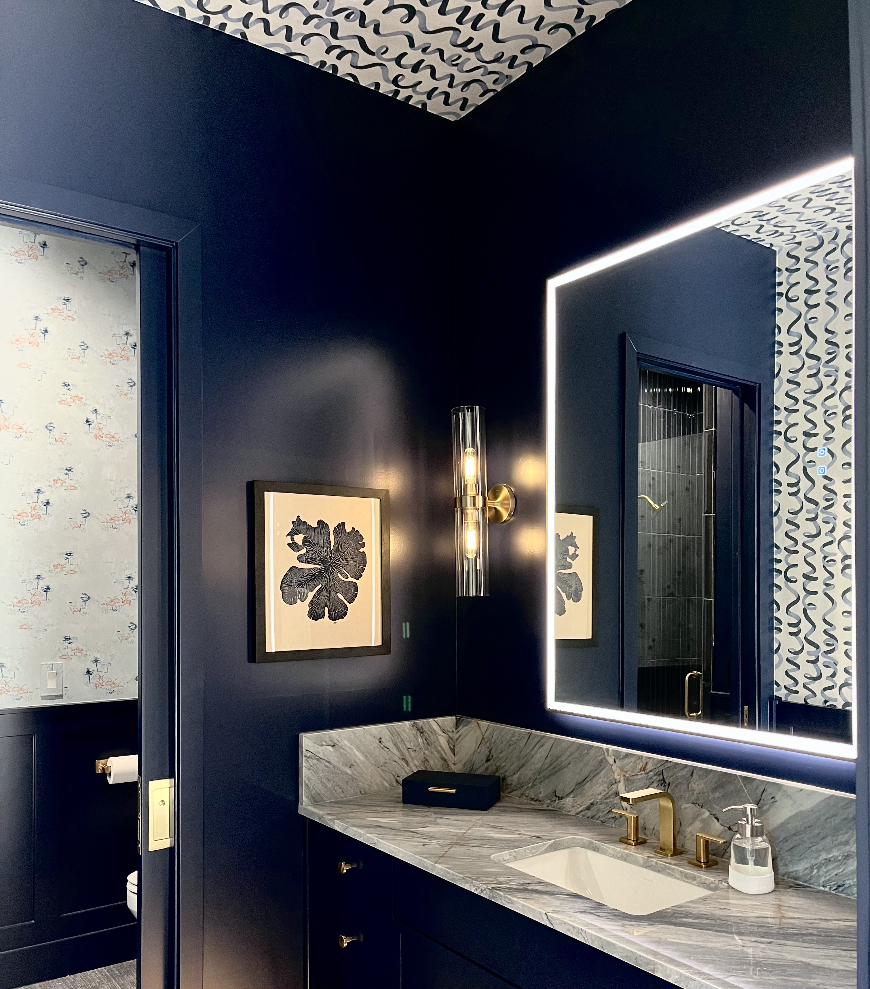 Navy powder room with squiggle-pattern ceiling wallpaper, marble vanity, and backlit mirror
